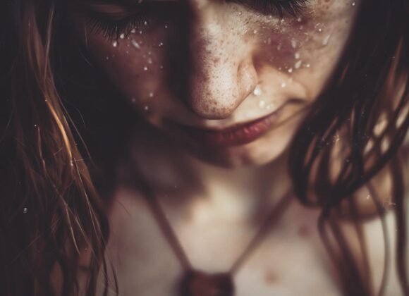 Close-up of a person with freckled skin looking down, with water droplets on their face and hair. They are wearing a necklace with a heart-shaped pendant, highlighting the importance of skin care and stress management in maintaining healthy, radiant skin.