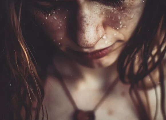 Close-up of a person with freckled skin looking down, with water droplets on their face and hair. They are wearing a necklace with a heart-shaped pendant, highlighting the importance of skin care and stress management in maintaining healthy, radiant skin.