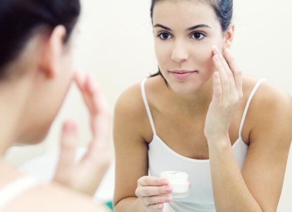 Effective Moisturizing Techniques for Eczema-Prone Skin A woman applies cream to her face while looking in a mirror. She holds a small jar of cream in one hand and uses the other hand to dab the cream on her cheek.
