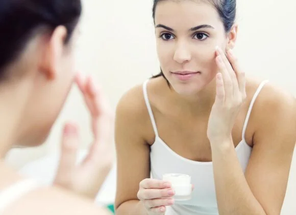 A woman applies cream to her face while looking in a mirror. She holds a small jar of cream in one hand and uses the other hand to dab the cream on her cheek.