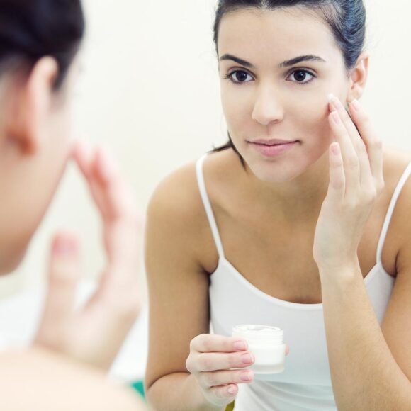 A woman applies cream to her face while looking in a mirror. She holds a small jar of cream in one hand and uses the other hand to dab the cream on her cheek.