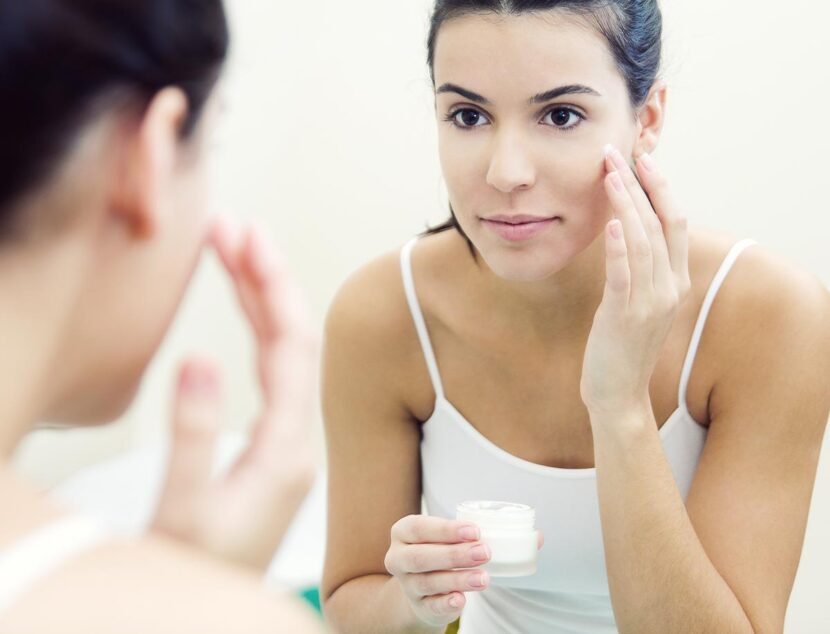 Effective Moisturizing Techniques for Eczema-Prone Skin A woman applies cream to her face while looking in a mirror. She holds a small jar of cream in one hand and uses the other hand to dab the cream on her cheek.
