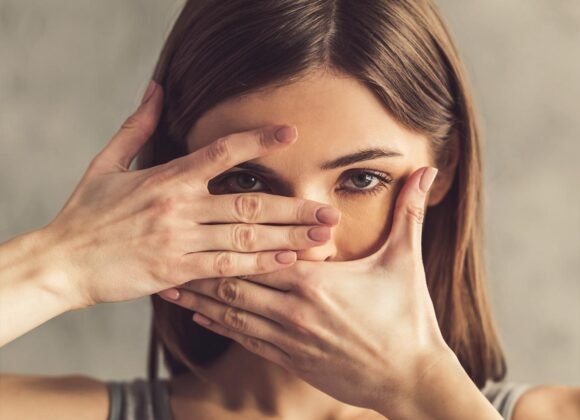 Facial Eczema: Symptoms and Skin Care Tips for Sensitive Skin A woman with brown hair frames her face with her hands, covering her mouth with one hand and peeking through a slit formed by her fingers of the other hand.