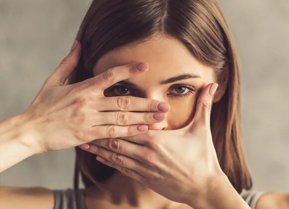 A woman with brown hair frames her face with her hands, covering her mouth with one hand and peeking through a slit formed by her fingers of the other hand.
