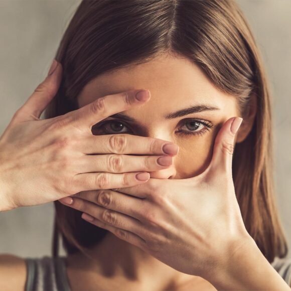 A woman with brown hair frames her face with her hands, covering her mouth with one hand and peeking through a slit formed by her fingers of the other hand.