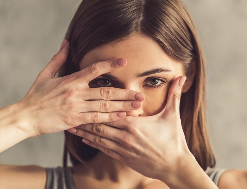 Facial Eczema: Symptoms and Skin Care Tips for Sensitive Skin A woman with brown hair frames her face with her hands, covering her mouth with one hand and peeking through a slit formed by her fingers of the other hand.