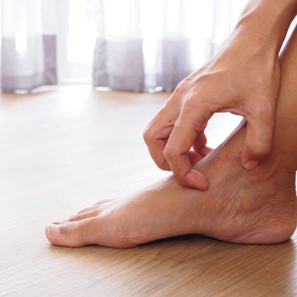A person scratches the inner side of their right ankle, struggling with dry patches, while seated on a wooden floor near a window with sheer curtains.