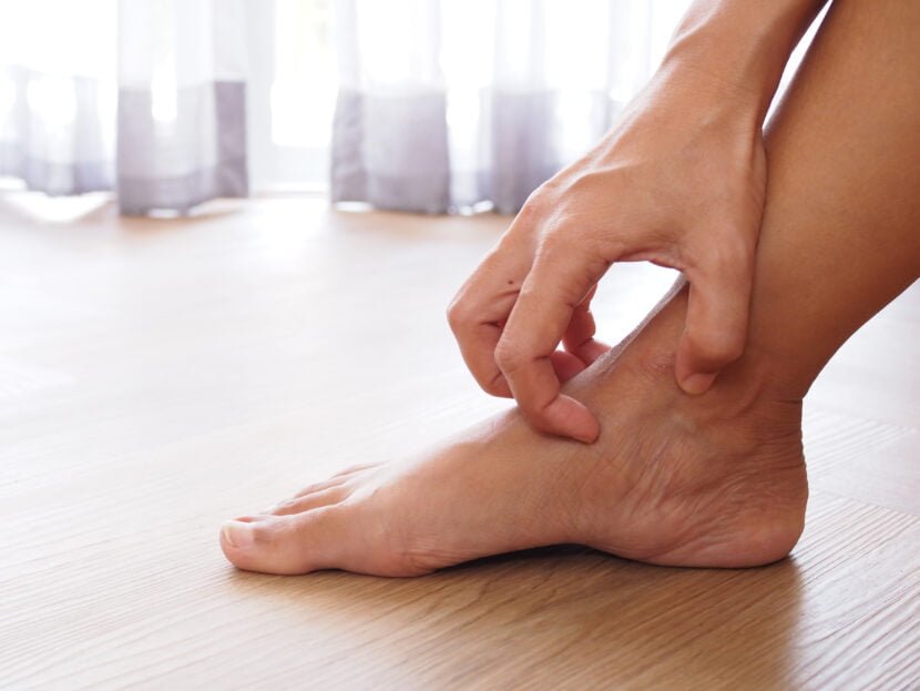 Foot Eczema: Causes, Symptoms, and Footwear Tips A person scratches the inner side of their right ankle, struggling with dry patches, while seated on a wooden floor near a window with sheer curtains.