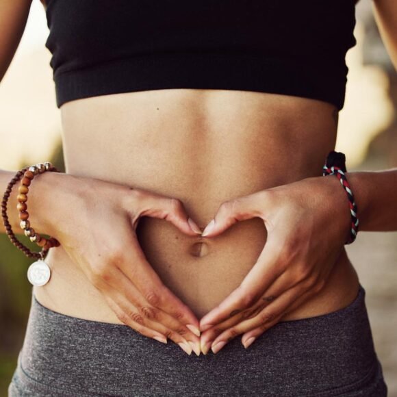 Person wearing a black crop top and bracelets, forming a heart shape with their hands over their stomach.