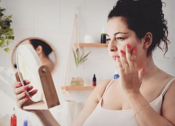 A woman with her hair up examines her skin, looking for signs of irritation or eczema triggers, while gazing into a handheld mirror in a bathroom with shelves and plants in the background.