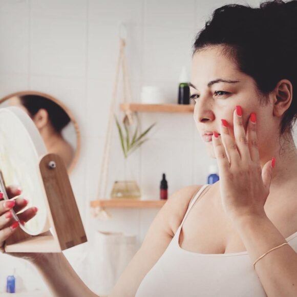 A woman with her hair up examines her skin, looking for signs of irritation or eczema triggers, while gazing into a handheld mirror in a bathroom with shelves and plants in the background.