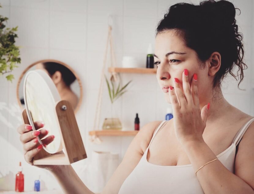 How Hormonal Changes Can Worsen Eczema Symptoms A woman with her hair up examines her skin, looking for signs of irritation or eczema triggers, while gazing into a handheld mirror in a bathroom with shelves and plants in the background.