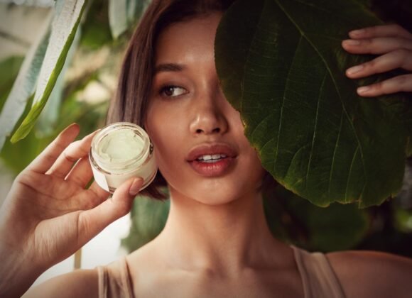 Natural Remedies for Eczema Symptoms That Actually Work A person holds a jar of cream next to their face, with one eye peeking out from behind a large green leaf.