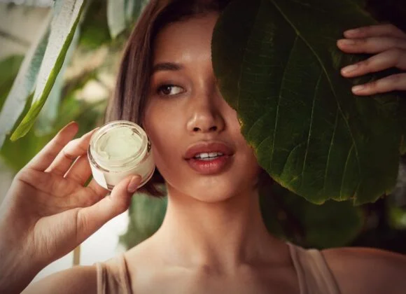 A person holds a jar of cream next to their face, with one eye peeking out from behind a large green leaf.