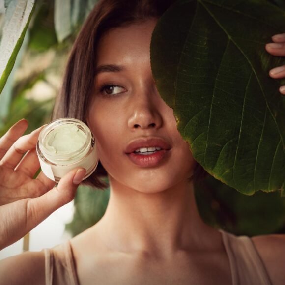 A person holds a jar of cream next to their face, with one eye peeking out from behind a large green leaf.
