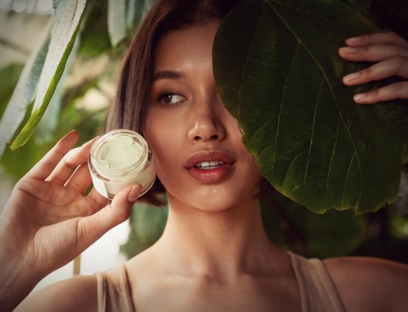 Natural Remedies for Eczema Symptoms That Actually Work A person holds a jar of cream next to their face, with one eye peeking out from behind a large green leaf.