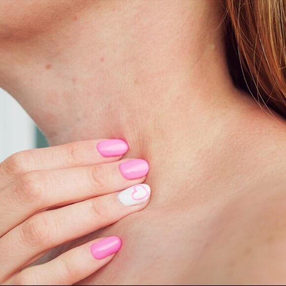 Close-up of a person touching their neck, with pink manicured nails and a heart design on the ring finger, possibly due to itchy skin.