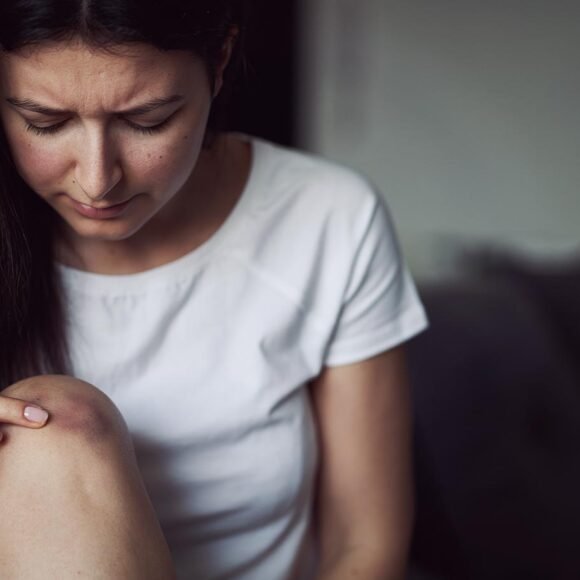 A woman with long dark hair wearing a white shirt looks at a bruise on her knee.