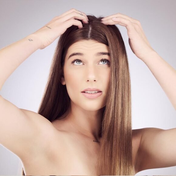 A woman with long straight hair lifts sections of her hair with both hands while looking upwards. She has a neutral expression, dry patches on her arms, and a small tattoo on her left wrist.