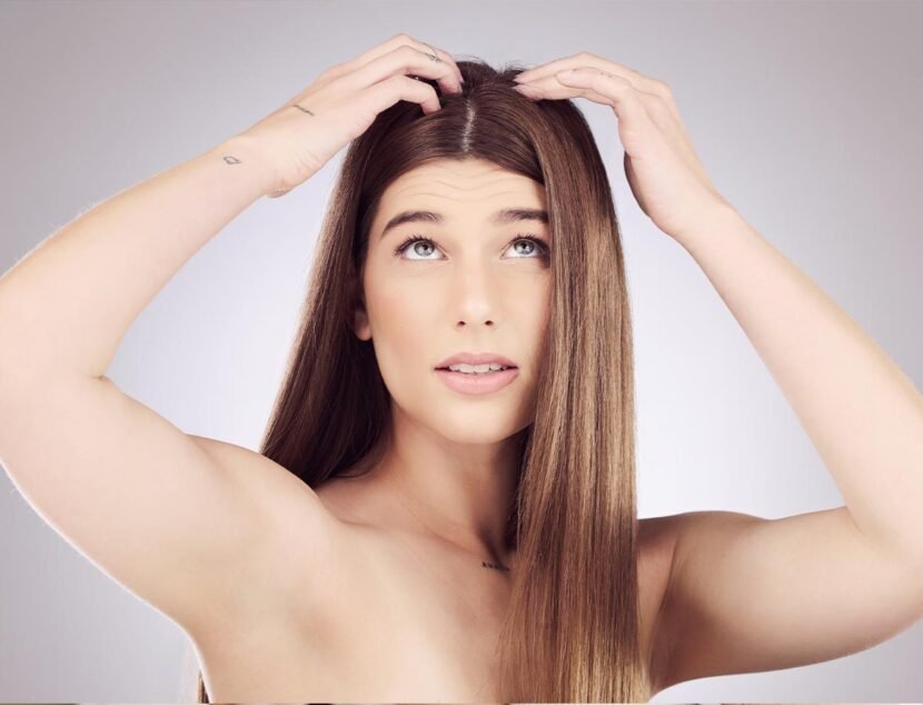 Scalp Eczema: Identifying Symptoms and Hair Health Impacts A woman with long straight hair lifts sections of her hair with both hands while looking upwards. She has a neutral expression, dry patches on her arms, and a small tattoo on her left wrist.